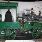 Image: A green automobile with a steam engine and spoked wagon wheels on display in a museum