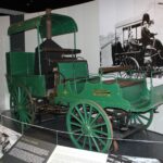 Image: A green automobile with a steam engine and spoked wagon wheels on display in a museum