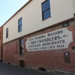 Image: A two-storey building with brick first floor and corrugated metal second floor. The words ‘Sail Makers, Riggers, Ship Chandlers. Provision Merchants Ship Stores in Bond Duty Paid’ are painted on a sign attached to the building’s first floor