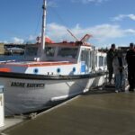 Image: Two men and a woman stand on a dock next to an historic motor vessel. The boat is white with an orange foredeck and cabin roof