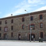 Image: A large, three-storey nineteenth century stone warehouse located next to a paved road. A sign in one of the windows reads ‘Est 1870, William Russell Pty Ltd., Sailmaker & Ship Chandler’