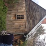 Image: A small, single-storey wooden dwelling with one visible window at a steep roof. The structure is surrounded by trees and vegetation