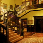 Image: An ornate wooden staircase in an historic house. Several ornate rugs and framed paintings are visible on the floors and walls surrounding the staircase