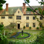 Image: The front of a large, two-storey stone mansion. A garden with benches and statuary is in the middle-foreground