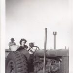 A woman and a small child sitting behind the steering wheel of a tractor.  A man behind them is sewing up a hessian bag.