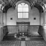 Image: the interior of a large hall with stone and wood panneled walls and a decorative wooden ceiling. The rear wall features a large window underneath which is a stage area with rows of chairs upon it and more chairs facing it.