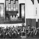 Image: an orchestra plays on a stage in front of a large pipe organ. The organ has two balcony areas within it upon which stand a man and a woman in formal dress.