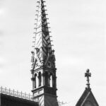 Image: a square gothic style open tower with a very steeply pitched roof rises from the centre of a crossed gable roof