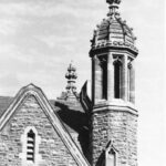 Image: a stone tower with a decorative domed roof rises above a simpler pitched slate roof.