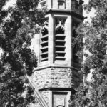 Image: A stone tower with a crenellated top can be seen between the branches of two trees. The tower is hexagonal in shape and features the Adelaide University coat of arms of the southern cross over an open book.