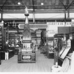 Image: Glass cases inside a building containing a variety of exhibits, and advertising signage, a man stands to the left dressed in uniform