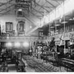 Image: Interior view of building overlooking a large hall with rows of seating, art exhibits, and glass cases