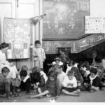 Image: a caucasian woman sits on a stool surrounded by children in a classroom with blackboards on the walls