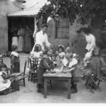 Image: two caucasian women teach small children to scrub tables and dust chairs outside of a kindergarten