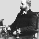 Image: black and white photograph of a seated man, dressed in black and holding a cane