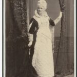 Image: Woman, dressed as the Queen, standing holding a curtain