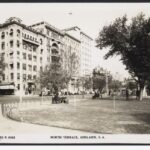 Image: Black and white photograph taken from across North Terrace looking West, with the Liberal Union Club in the distance. The photograph was taken in the 1950s and shows people sitting on park benches looking towards the Liberal Union Club building.