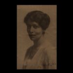 Image: A photographic head-and-shoulders portrait of a young Caucasian woman dressed in early twentieth century attire. She has dark hair and eyes and is wearing a pearl necklace and light cardigan sweater