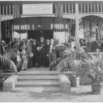 Image: a group of men and women in early 20th century dress sit or stand in front of a doorway in the shade of a verandah decorated by flags. Palms in pots next to the stairs leading to the verandah dominate the foreground.