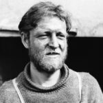 Image: A bearded young man wearing a heavy wool sweater stands outside a hut. His face shows evidence of frostbite