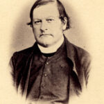 Image: A photographic head-and-shoulders portrait of a Caucasian man in Victorian-era priest’s attire