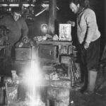 Image: Two men stand in a darkened room next to a pot-bellied stove. One observes the other as he welds a metal vise