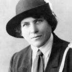 Image: A photographic head-and-shoulders portrait of a young Caucasian woman dressed in an early twentieth century Girl Guides uniform with hat. She has light-coloured eyes and dark hair