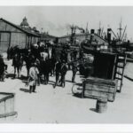 Image: A group of men mill about on a wharf. Two steamships are visible in the background. What appears to be a horse-drawn cart tipped on its side is located at the edge of the wharf