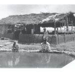 Image: Three men crouch near a pool next to a small, thatch-roofed structure in a remote area