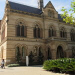 Image: a two storey gothic style building made of a yellow toned stone with darker horizontal stripes. A porch/balcony protrudes from the front. The ground floor windows are simple arches and set in pairs but the upper floor windows are of a church style