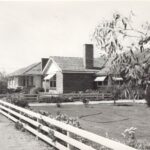 Image: A row of modern brick homes with well-manicured lawns and white fences are located next to a dirt road