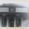 Image: An Islamic symbol of a crescent moon and star is painted just below the roof on a building fronted with arches. A man bends down behind a narrow column with one leg bent at the knee, possibly removing or putting on a shoe.