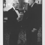 Image: Two men in tuxedos stand face to face. The man on the right has his hand raised, one finger pointed up, gesturing. The other, who holds a piece of paper in his hand, is smiling.