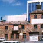 Image: Colour photograph of an historic bluestone building with red brick quoins and detailing surrounded by sky scrapers and fronted by a carpark