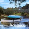 Image: a postcard with the greeting "Greetings from Adelaide". The image is of a small blue and white boat with a covered seating area sailing down a wide river which winds through parklands. In the background is a green metal footbridge.