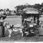 Image: black and white shot of a young girl with a goat toting a decorated cart of produce, with a toddler holding a bottle behind the cart