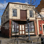 Image: A two-storey building with brick first floor and corrugated metal second floor. The words ‘Hy. Weman, Sail Maker, Ship Chandler’ are painted on the building shop front