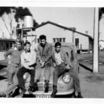 Image: four young men sitting on bonnet of car, buildings in background