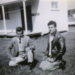 Image: two men kneeling on grass in front of buildings