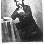 Image: Black and white photograph of a man seated backwards on chair with arms crossed on seat back and an open book on the table beside him