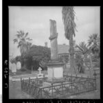 Image: A tall broken stone column on top of a large stone base inscribed with names. A short fence surrounds the memorial.