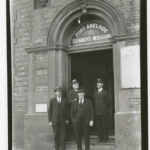 Image: Four Caucasian men in early twentieth century attire stand outside the front entrance of a large bluestone building. A sign above the entrance door reads ‘Port Adelaide Seamen’s Mission’