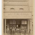 Image: A group of Caucasian men in late nineteenth-century labourer attire stand in front of a shop. The words ‘Jas. R. McColl, House and Ship Plumber, Tinsmith, &c.’ are painted on the front of the building