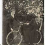 Image: a woman sitting astride a bicycle, with two younger women alongside