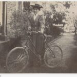 Image: a woman standing beside a bicycle at the side of a stone building