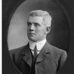 Image: A photographic head-and-shoulders portrait of a young Caucasian man wearing an early Edwardian suit. He is clean shaven and has light-coloured hair and eyes