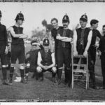 Image: A group of Australian Rules Football players in early twentieth century uniforms pose for a photograph. Most of the men are wearing period police helmets