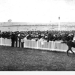 Image: Black and white photo of horseracing in progress, crowd behind a fence