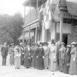 Image: A group of women and men in Edwardian attire stand in front of a two-storey stone building with flags hanging from the second-floor verandah