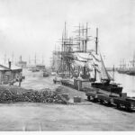 Image: Several late nineteenth century sailing ships are moored alongside a large wharf in a port town. A large pile of stone ballast and rail cars are visible in the foreground, while a single-storey bluestone building is located nearby